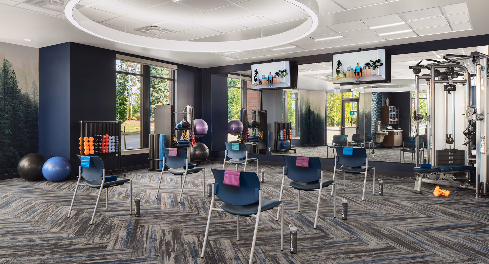 A senior living facility exercise room with six chairs spaced apart, each with a towel draped over the back and a water bottle on the floor nearby. The room has large windows, mirrors on one wall, exercise balls, dumbbells, and a multi-function gym machine. Two TVs mounted on the wall display an exercise video with a person demonstrating seated exercises.