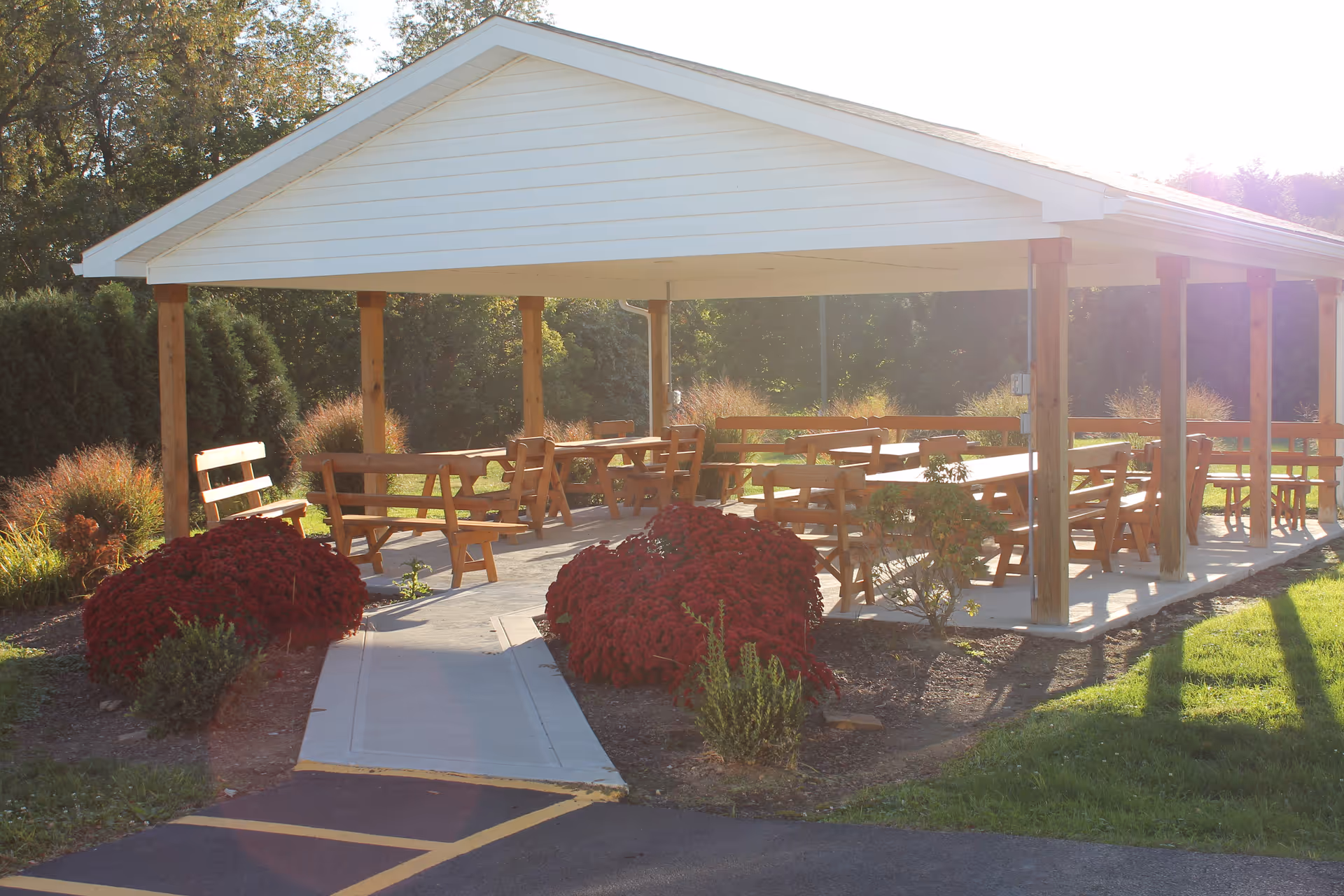 Outdoor covered pavilion with wooden benches and tables surrounded by landscaped bushes and greenery, with sunlight filtering through trees in the background.