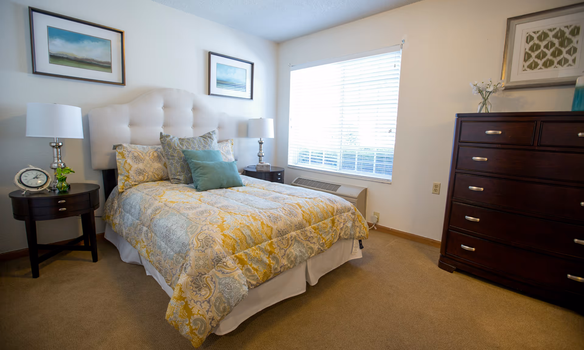 Bright furnished bedroom with a bed, nightstands and lamps, a large window, and a dark wood dresser.