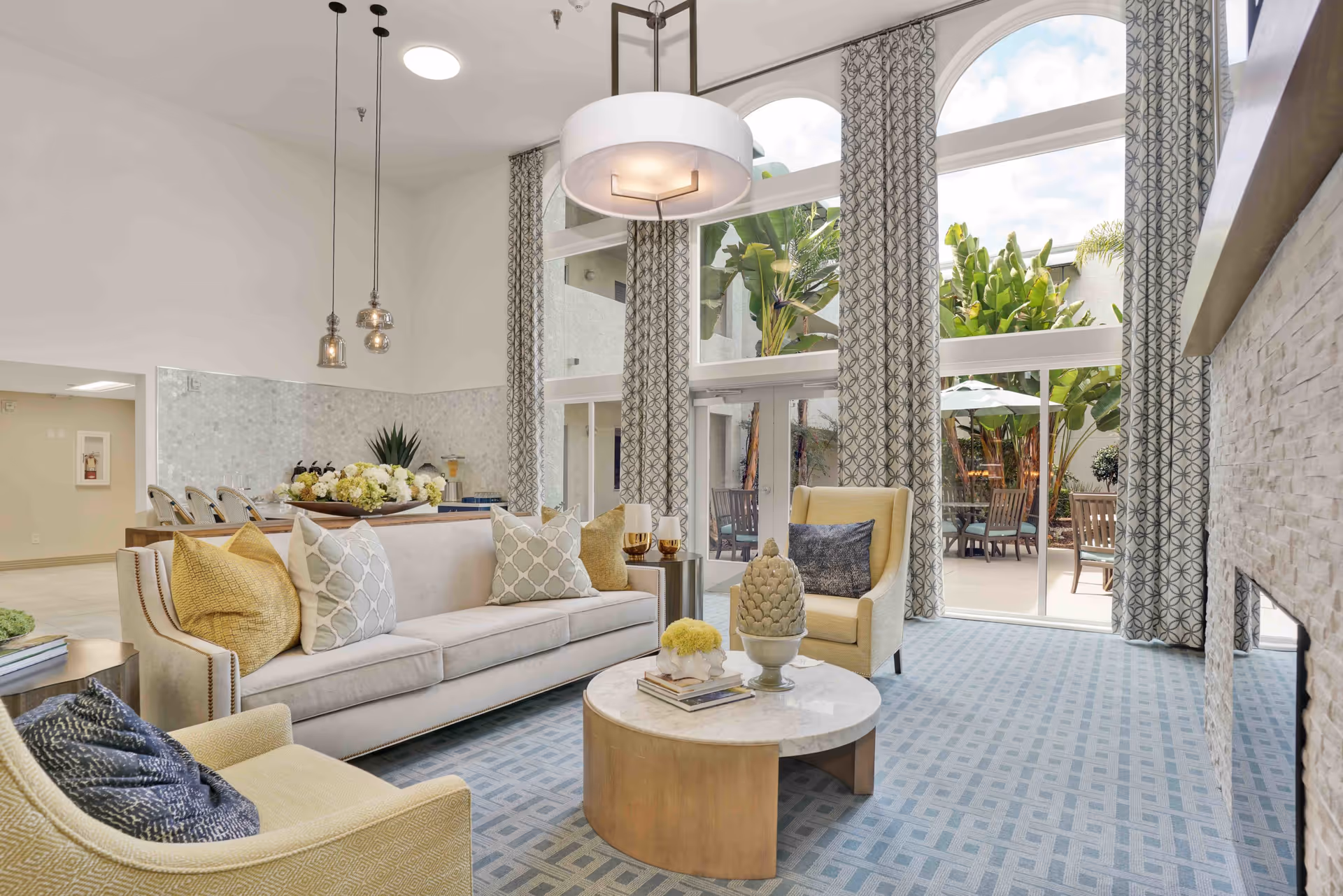 Bright and spacious senior living facility common area with a beige sofa adorned with patterned pillows, two armchairs, a round coffee table with decorative items, large windows with patterned curtains letting in natural light, and a view of an outdoor patio with chairs and greenery.