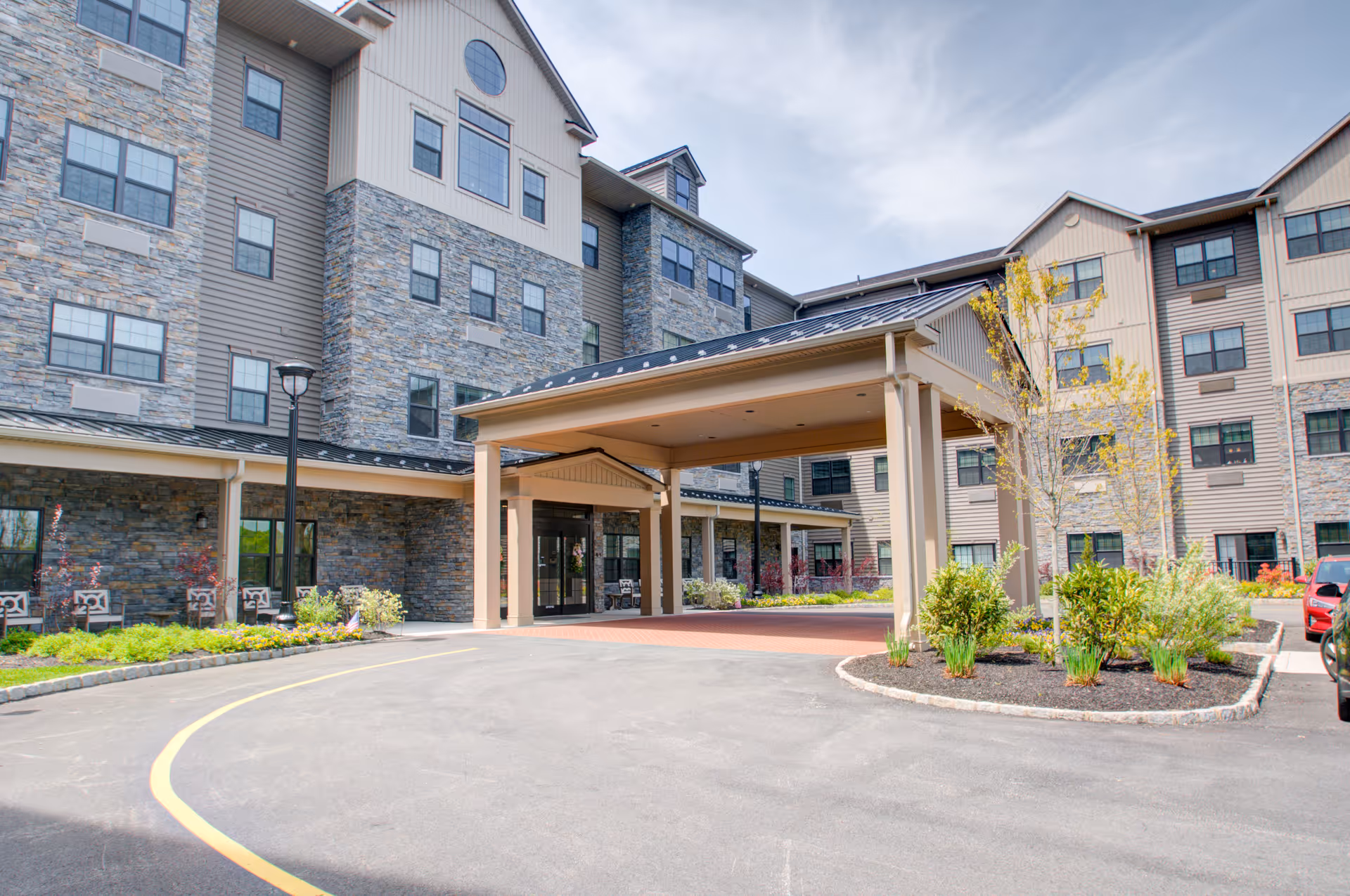 Exterior view of The Pinnacle at Plymouth Meeting senior living facility showing a multi-story building with stone and siding facade, large covered entrance, landscaped greenery, and a paved driveway.
