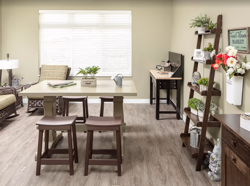 A bright and cozy room with a large window covered by white blinds. In the center is a rectangular table with four wooden stools around it. On the table are a small watering can, a plant in a wooden box labeled 'GARDEN', and some books. To the left, there are cushioned wicker chairs and a floor lamp. On the right side, there is a wooden ladder shelf with various potted plants, decorative items, and a wall-mounted container holding flowers. A small workbench with tools and supplies is against the far wall. The floor is light wood, and the walls are painted a soft beige.