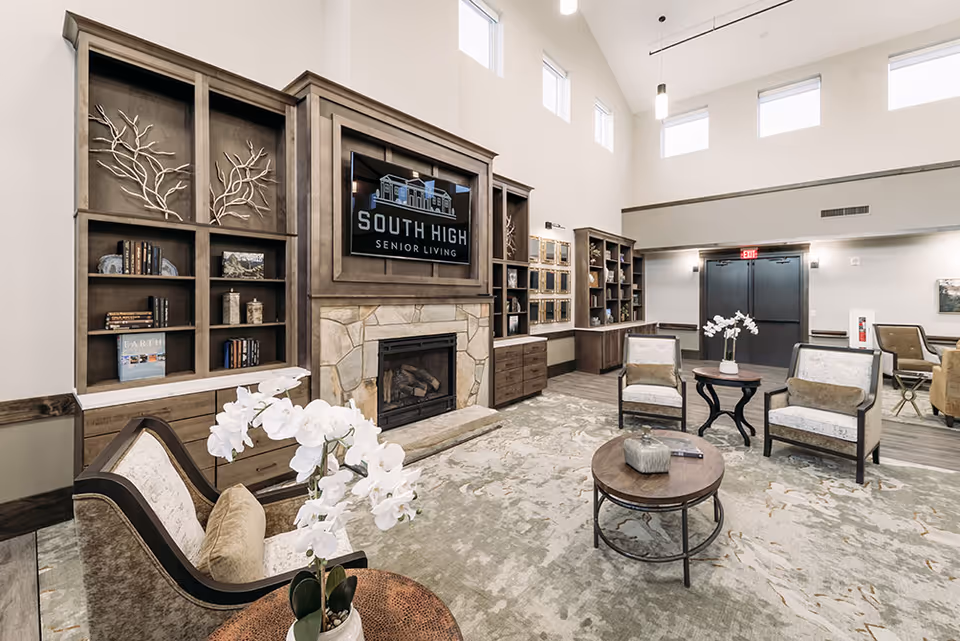 A spacious and well-lit senior living facility common area with high ceilings and multiple windows. The room features a stone fireplace with a large TV above it displaying the South High Senior Living logo. Surrounding the fireplace are built-in wooden shelves with decorative items and books. Comfortable armchairs and round wooden tables with white orchid plants are arranged on a patterned carpet. The walls are painted in neutral tones, and there are double doors and additional seating visible in the background.