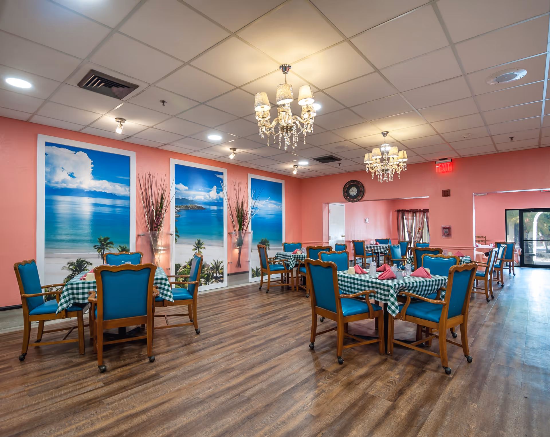 A senior living facility dining room with several tables covered in green and white checkered tablecloths, each set with pink napkins and silverware. The chairs have wooden frames with teal cushions. The walls are painted pink and decorated with large beach-themed murals and tall vases with decorative branches. The ceiling has recessed lighting and two ornate chandeliers. The floor is wood, and there is an exit door visible in the background.