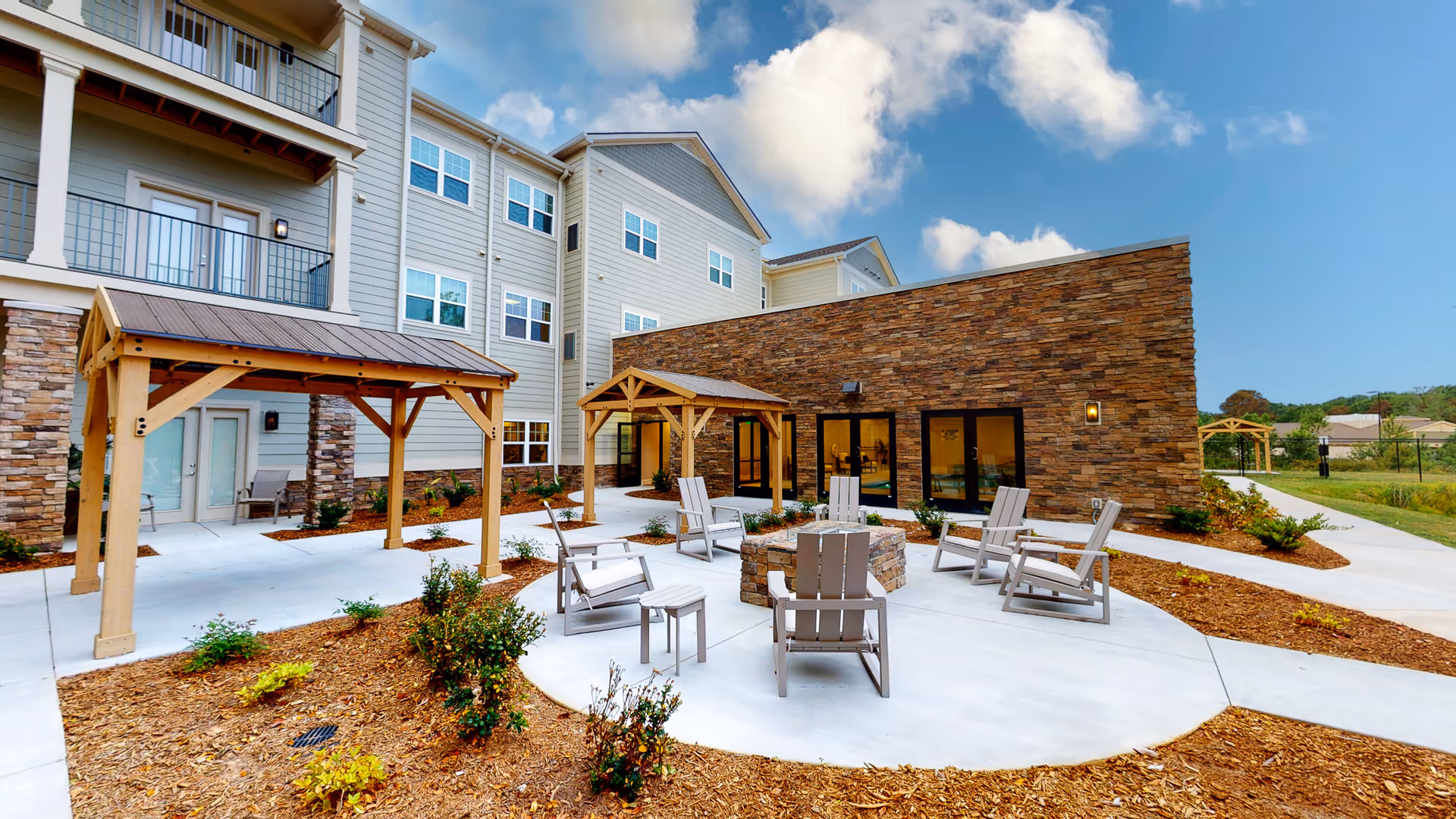 Outdoor patio area at Legacy Reserve at Fairview Park featuring a circular concrete seating area with Adirondack chairs around a fire pit, wooden pergolas, landscaped garden beds with small shrubs, and a multi-story building with balconies and large windows under a partly cloudy sky.