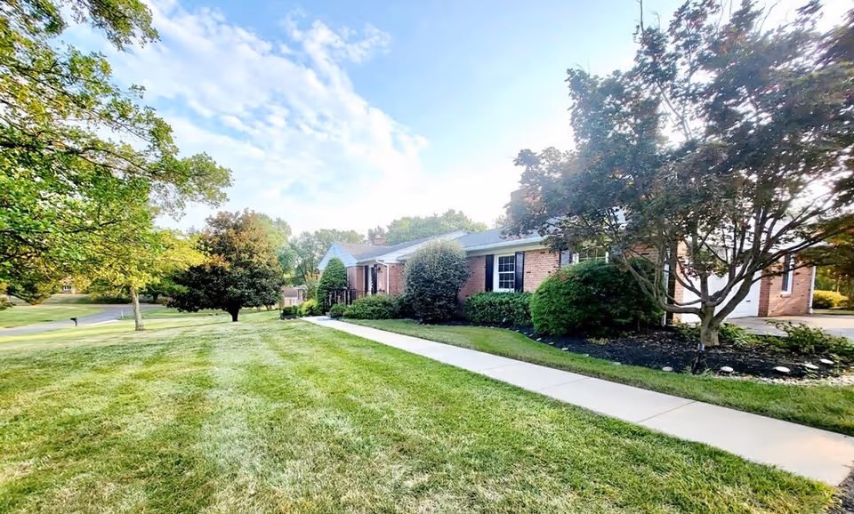 Front view of a single-story brick assisted living building with a sidewalk, manicured lawn, and trees.