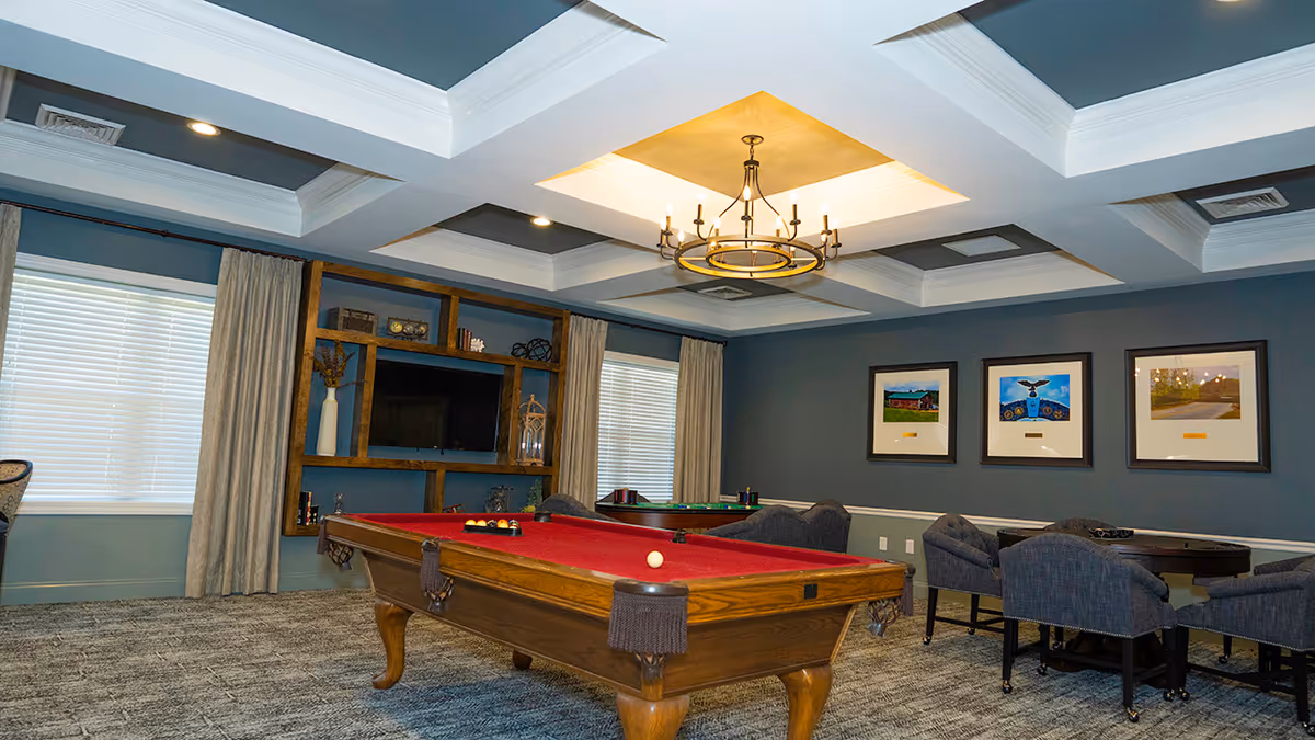 Game room with a red-felt pool table, armchairs and tables, TV shelving, and framed artwork beneath a coffered ceiling.