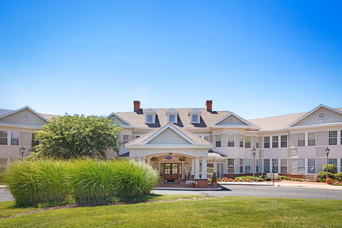 Front exterior of a large two-story senior living facility with a covered entrance, landscaped grounds, and a clear blue sky.