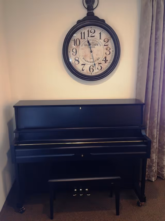 A black upright piano with a matching bench is positioned against a beige wall. Above the piano hangs a large round wall clock with a vintage design and black frame. To the right, beige curtains with a subtle pattern are partially visible.
