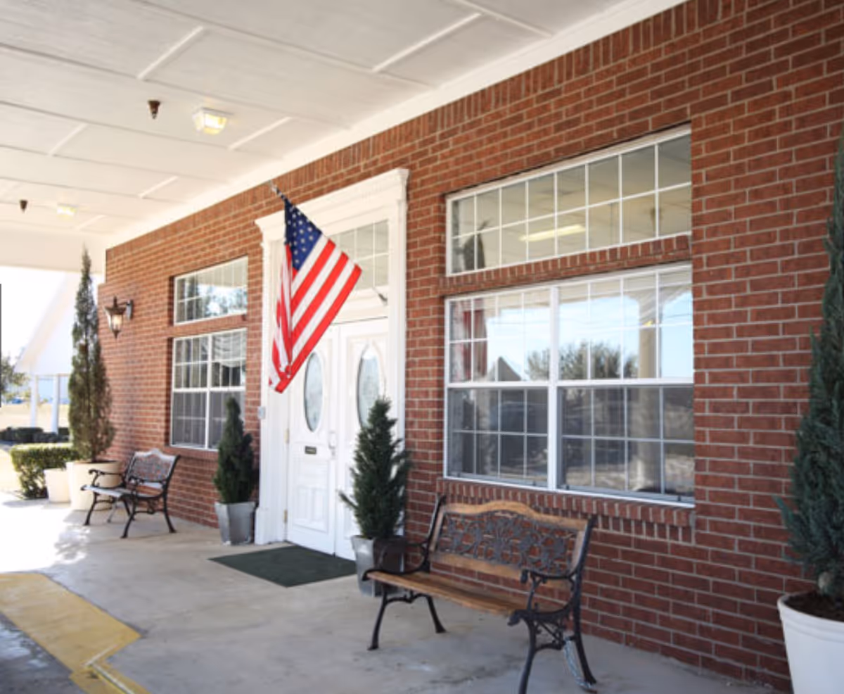 Covered entrance area of a brick building with white double doors, an American flag mounted above the doors, two benches, potted plants, and large windows.