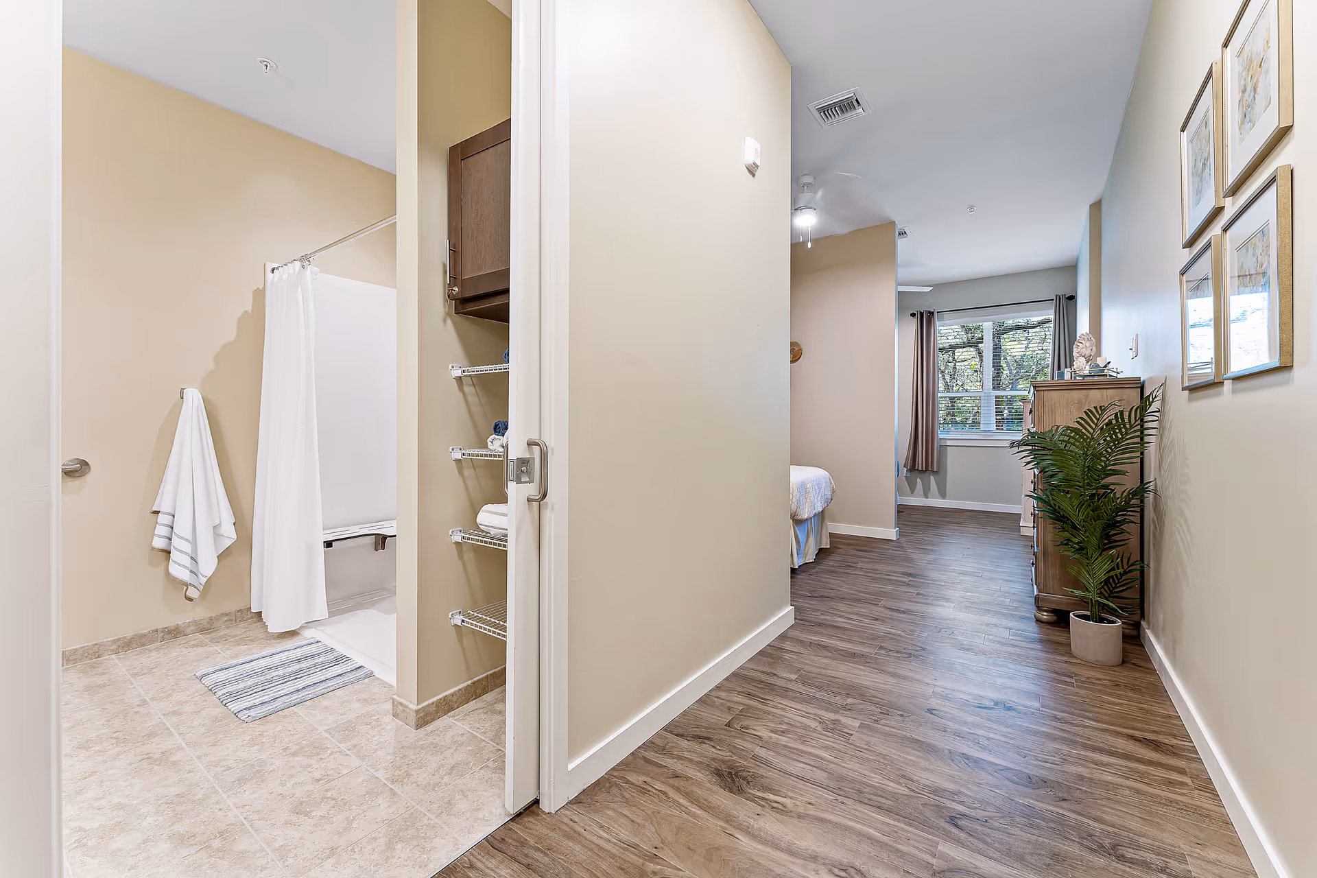 Interior hallway of an apartment showing an open bathroom on the left and a bedroom area at the far end with wood floors and a potted plant.