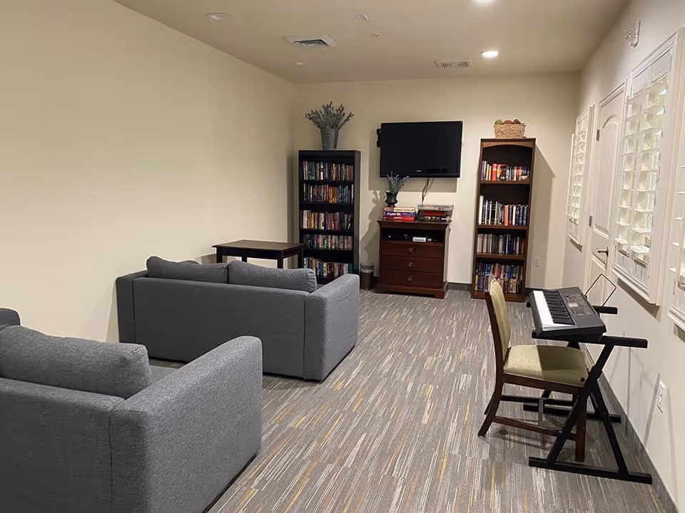 A cozy living room area with two gray sofas facing a wall-mounted flat screen TV. There are two tall bookshelves filled with books on either side of a wooden cabinet beneath the TV. A small table is placed behind one sofa. On the right side of the room, there is a keyboard on a stand with a chair in front of it. The room has light-colored walls and a patterned carpet floor.