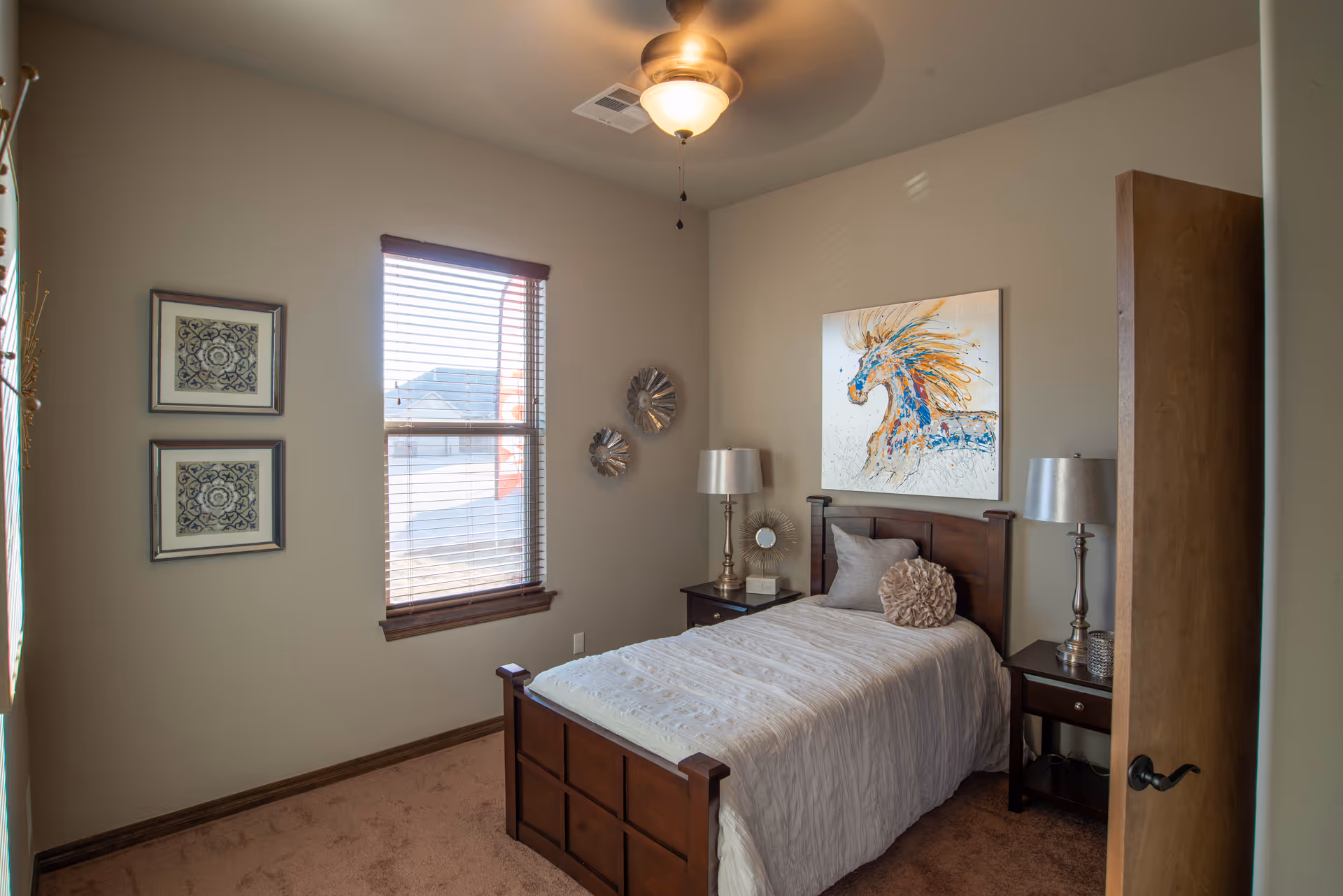 A small bedroom with a single wooden bed covered in a white comforter and two decorative pillows. There are two matching wooden nightstands on either side of the bed, each with a silver lamp. Above the bed is a colorful painting of a horse. The room has beige walls, a window with wooden blinds, two framed artworks on the left wall, and a ceiling fan with a light fixture.