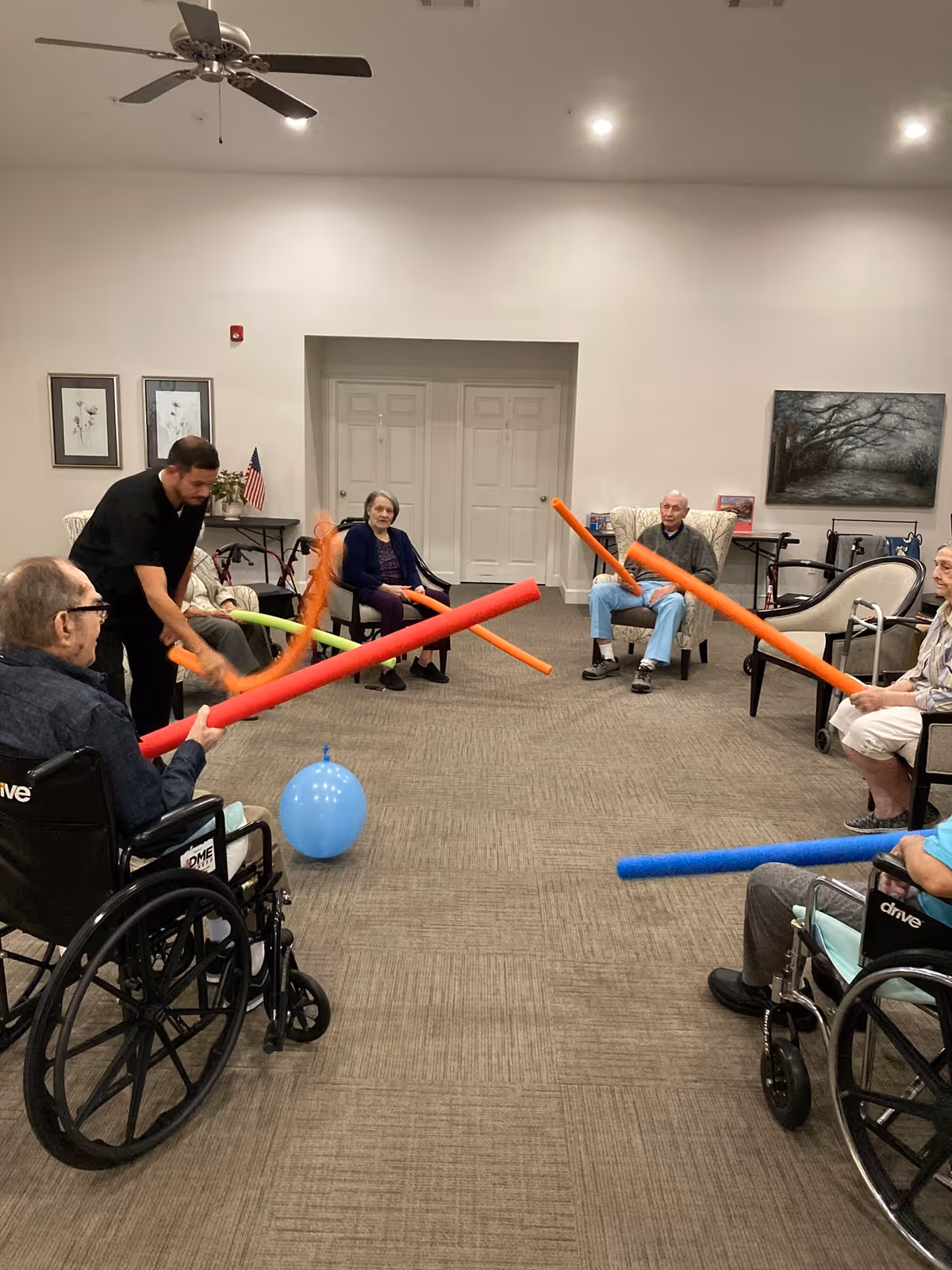 A group of elderly people and a caregiver in a common room of an assisted living facility. They are seated in a circle, some in wheelchairs, holding colorful foam noodles and playing with a blue balloon. The room has beige carpet, white walls, framed artwork, and a ceiling fan.