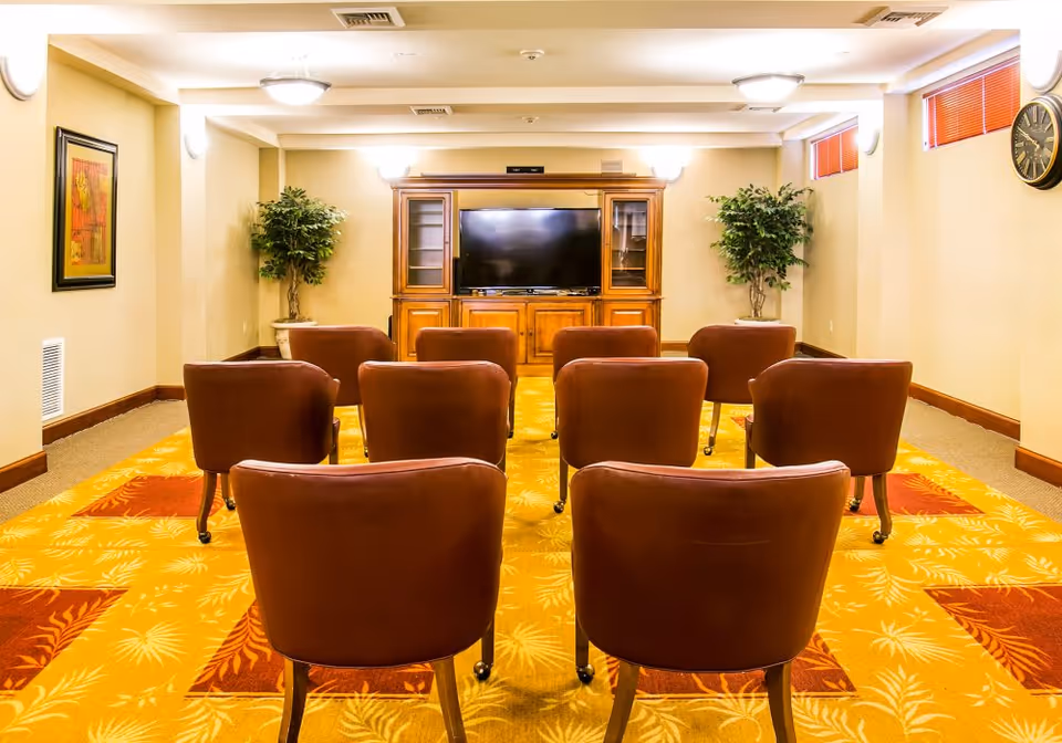 A small media room with eight brown leather chairs arranged in two rows facing a large flat-screen TV set in a wooden entertainment center. The room has beige walls, two potted plants on either side of the TV, a framed picture on the left wall, a clock on the right wall, and a patterned yellow and red carpet.