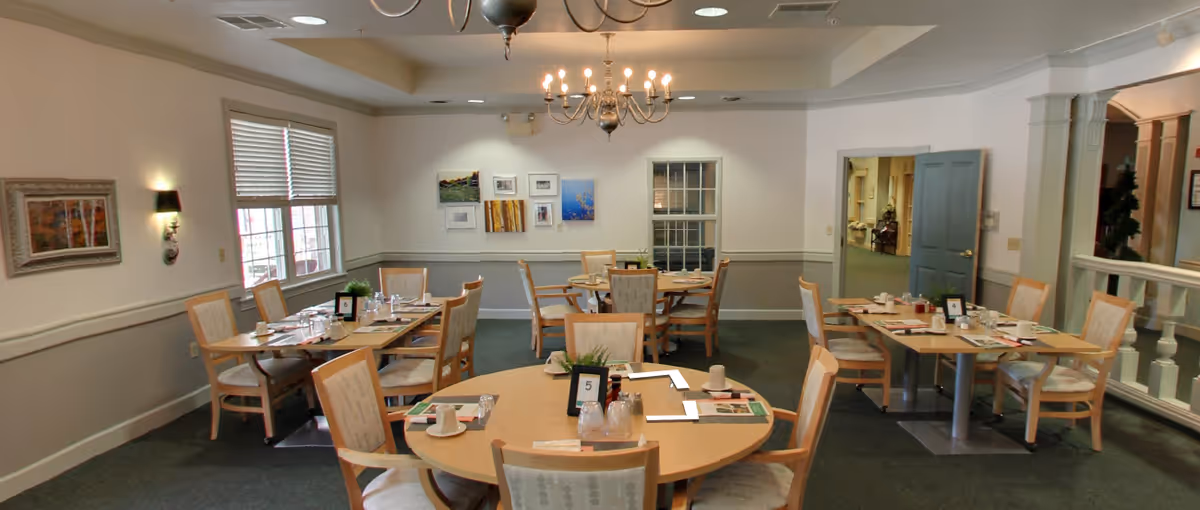 A dining room with several round and rectangular tables set with placemats, cups, and utensils. The room has beige walls with white trim, a chandelier hanging from the ceiling, framed artwork on the walls, and windows letting in natural light. There are wooden chairs with cushioned seats around each table.