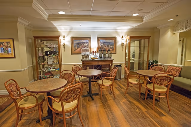 A cozy common area with wooden floors featuring three round tables surrounded by cushioned chairs with floral upholstery. Against the wall, there is a wooden sideboard with coffee dispensers and cups, flanked by two glass display cabinets. The walls are decorated with framed paintings and sconces providing warm lighting.