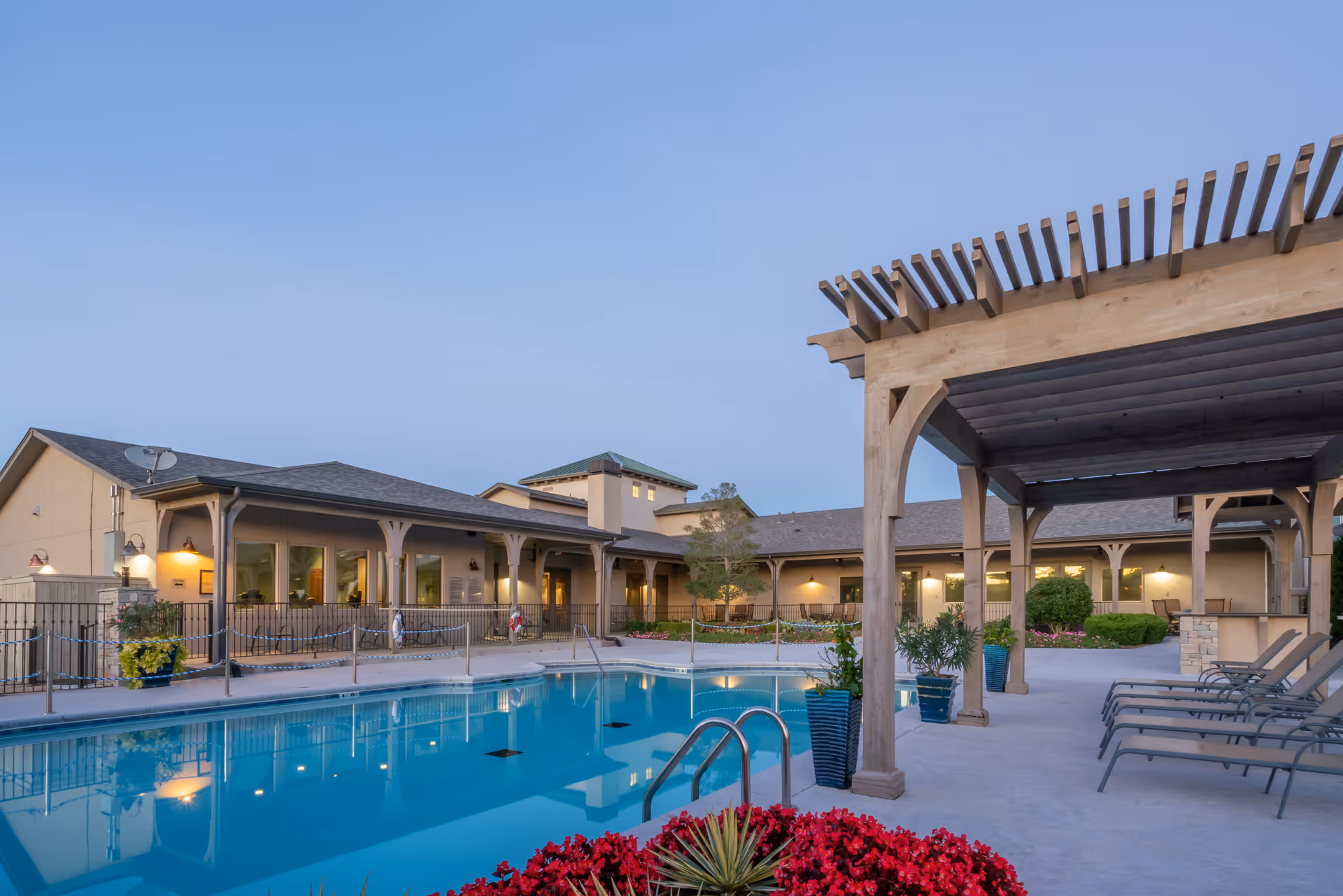 Outdoor swimming pool area at Hyde Park at Tulsa Hills with lounge chairs under a wooden pergola, potted plants, and a building with large windows in the background during early evening.