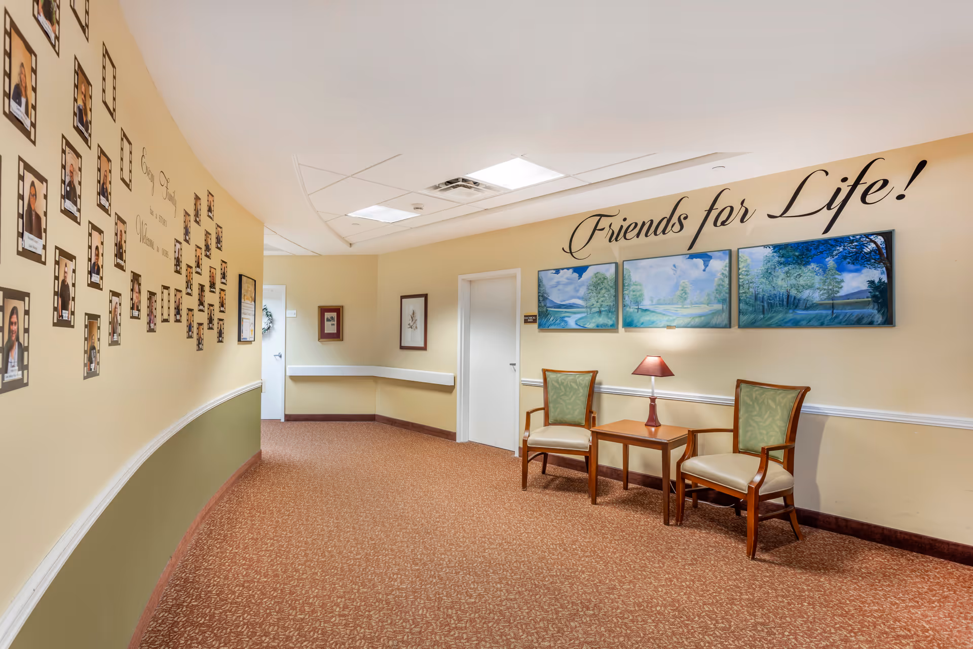 Carpeted hallway/common sitting area with two chairs and a table beneath wall art that reads 'Friends for Life!' and a wall of framed resident photos.