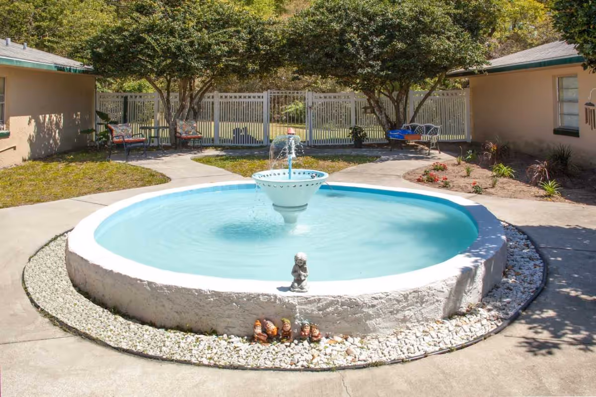 Outdoor courtyard area with a circular water fountain in the center surrounded by a white stone border. There are two beige buildings on either side with green-trimmed windows. The courtyard has concrete walkways, small garden beds with plants and flowers, and several seating areas with chairs and a bench under trees. A white fence with a gate is visible in the background.