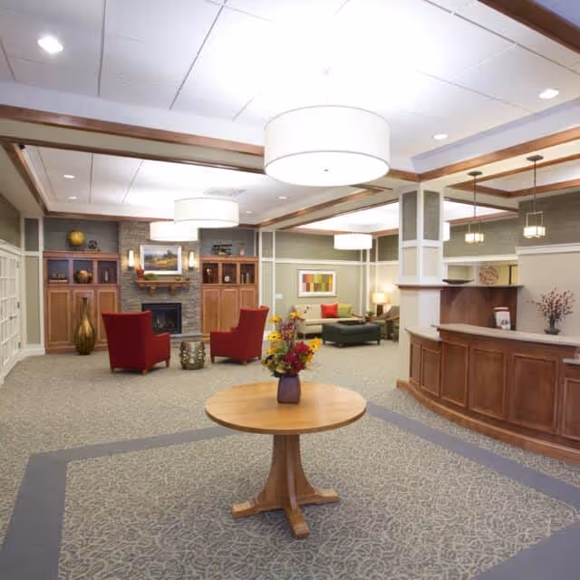 Bright assisted living lobby with a round center table holding flowers, seating areas, a fireplace, and a reception desk.