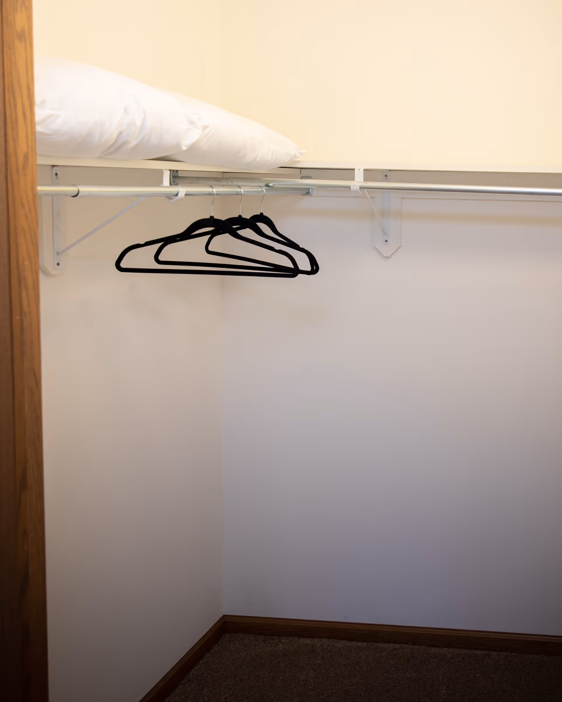 Empty closet with three black hangers hanging on a metal rod and a white pillow placed on the shelf above the rod. The closet has beige walls and brown carpeted floor.