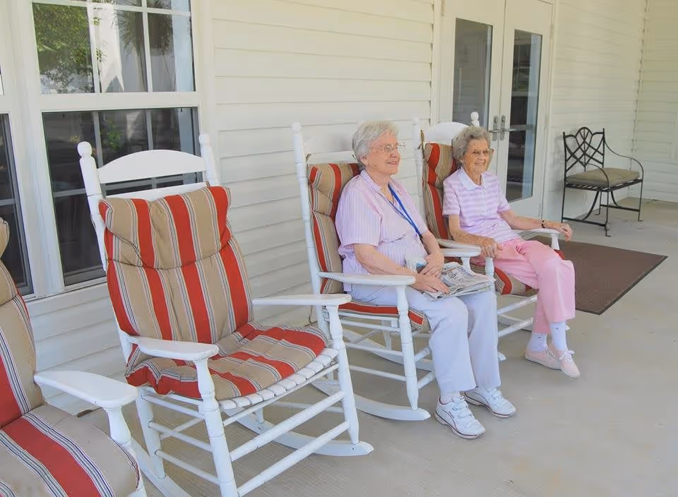 Two elderly women sitting and relaxing on white rocking chairs with red and beige striped cushions on a covered porch outside a building with white siding and windows.