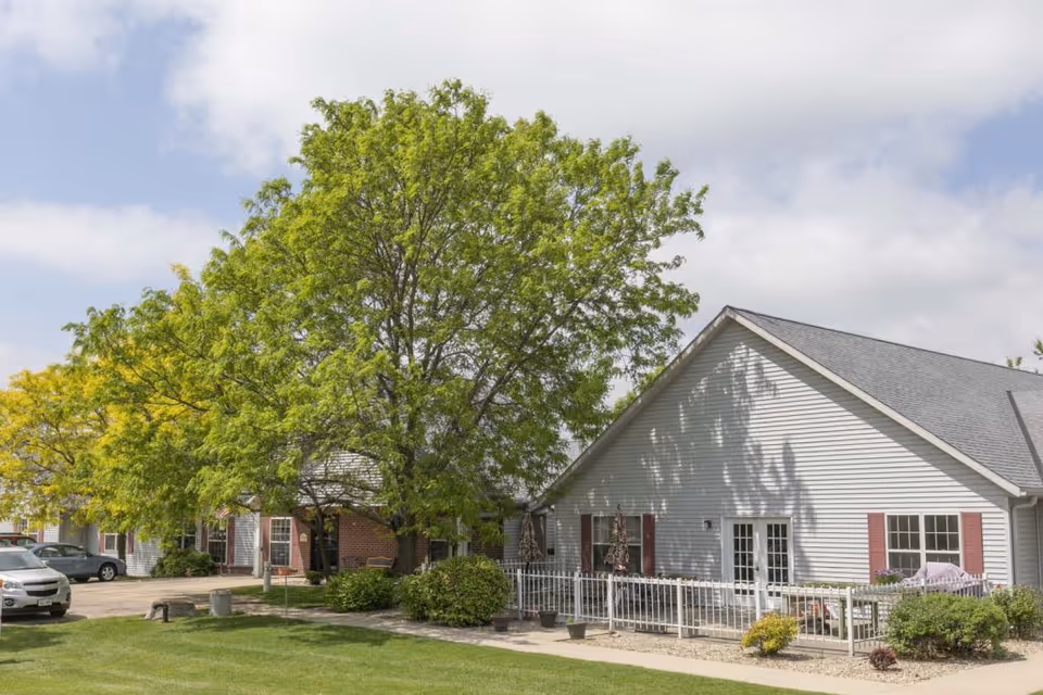 Front exterior of a single-story assisted living building with a large tree, lawn, and parked cars.