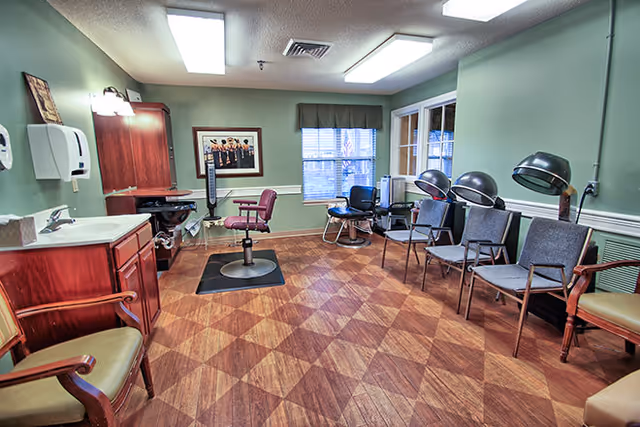 A salon-style interior with a barber chair, sink and cabinetry, several waiting chairs and hooded hair dryers under fluorescent lights.