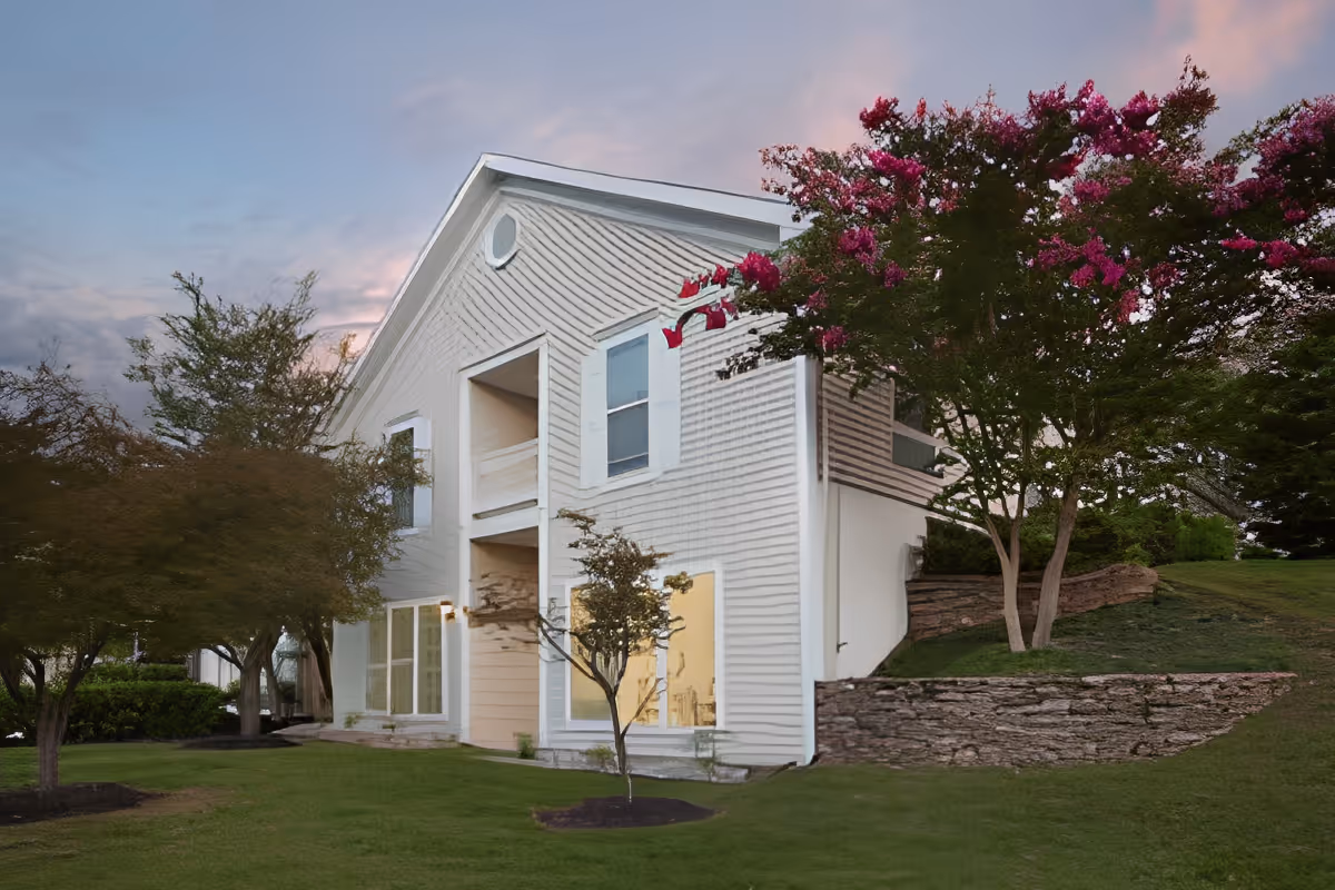 Exterior view of a two-story white building with large windows and a balcony, surrounded by green grass, trees, and a stone retaining wall under a partly cloudy sky at dusk.