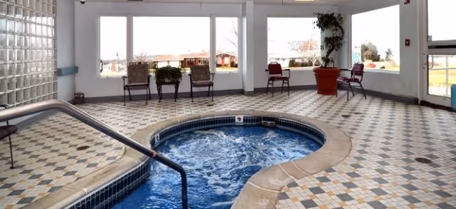 Indoor hot tub or small pool with bubbling water surrounded by tiled floor and large windows showing an outdoor view. Several chairs and potted plants are placed near the windows.