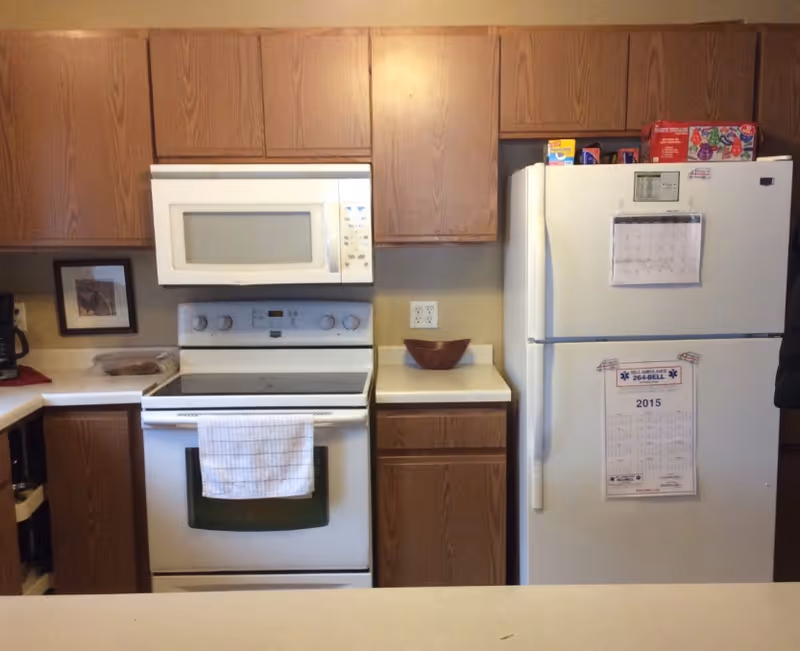 A kitchen with wooden cabinets, a white microwave above a white electric stove with a towel hanging on the oven door, and a white refrigerator with a calendar and notes attached to it. There is a brown bowl on the countertop next to the refrigerator and some food items on top of the fridge.