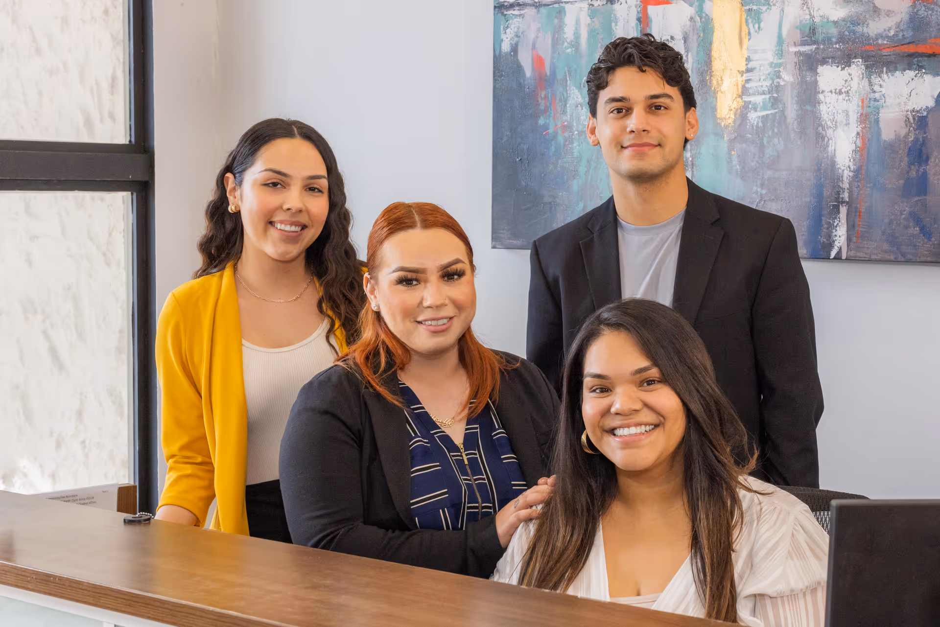 Four young adults smiling and posing behind a reception desk in a modern office setting with a large abstract painting on the wall behind them.
