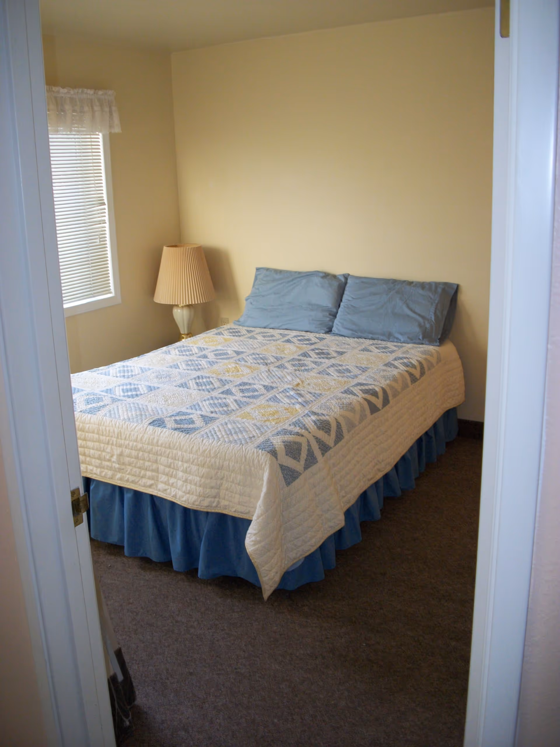 A small bedroom with a bed covered in a quilt with blue and yellow patterns, two blue pillows, a blue bed skirt, a beige wall, a window with blinds and a white lace valance, and a beige lamp on a small table in the corner.