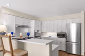 A modern kitchen with white cabinets, a stainless steel refrigerator, built-in oven, microwave, and a white countertop island with two chairs. The kitchen is well-lit with recessed ceiling lights and has a clean, minimalist design.