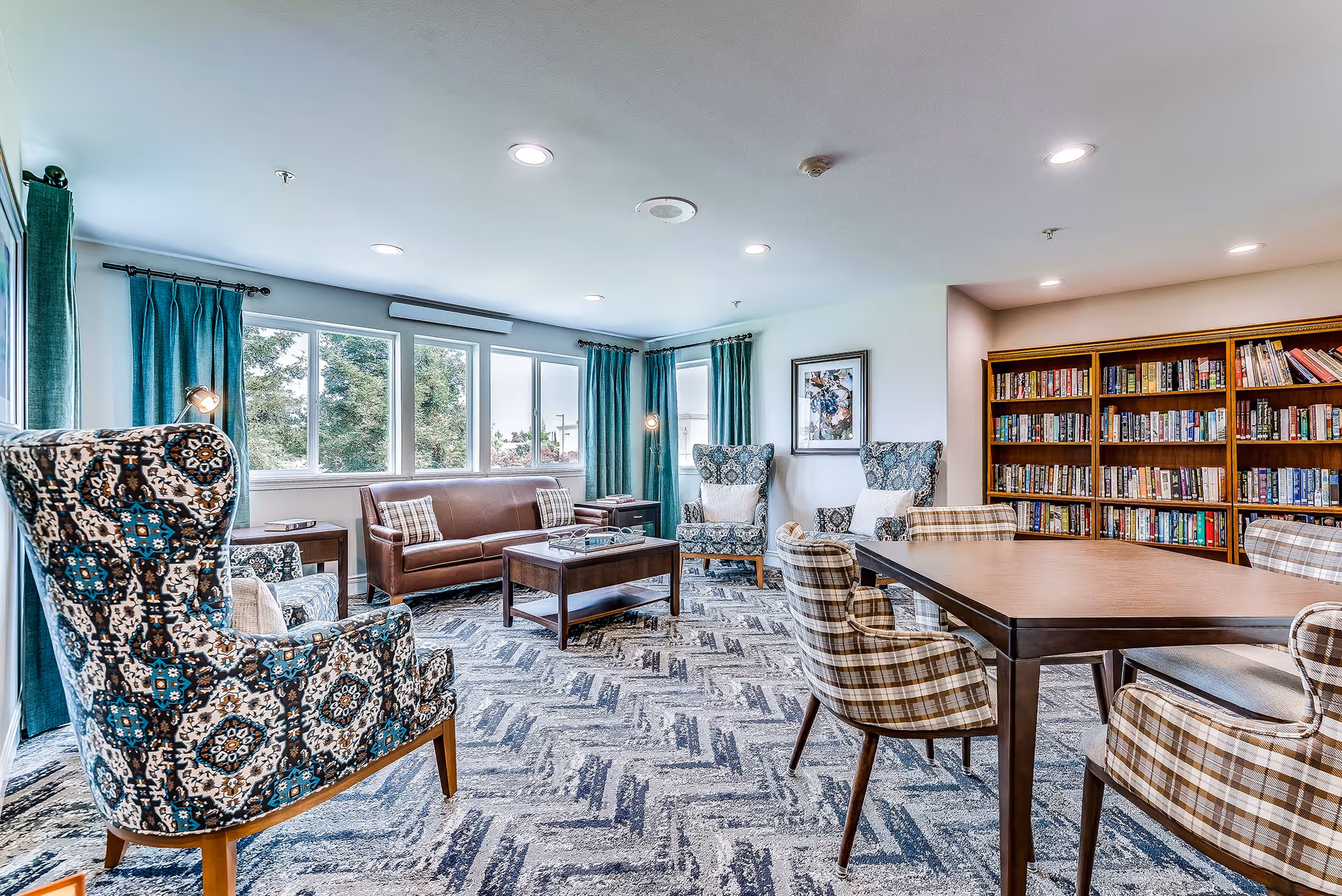 A bright senior living lounge with patterned armchairs, a sofa, a coffee table, a dining table with plaid chairs, bookshelves, and large windows with teal curtains.