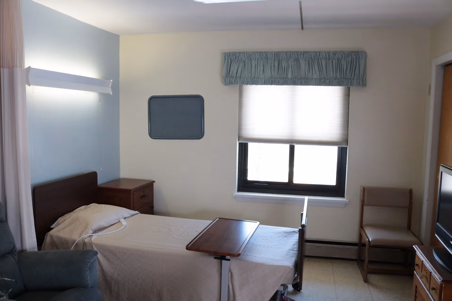 A simple, clean bedroom in a senior living facility with a single bed covered in a beige blanket, a wooden bedside table, a window with a blue valance and a white blind, a wooden chair, a TV on a wooden dresser, and a green armchair partially visible on the left.