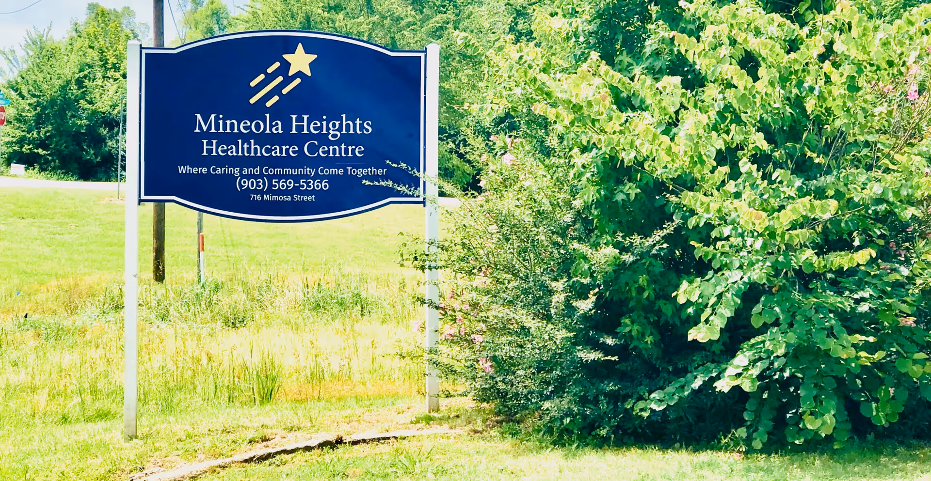 Blue roadside sign reading "Mineola Heights Healthcare Centre" on a grassy lawn beside dense green shrubs.