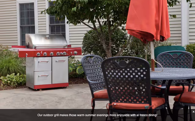 Outdoor patio with a red gas grill, round table and chairs with red cushions and a closed red umbrella in front of a building.