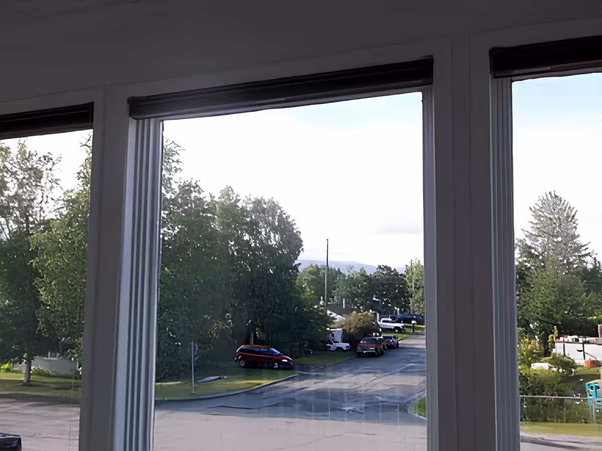 View through large windows showing a street with parked cars, green trees, and a partly cloudy sky outside.