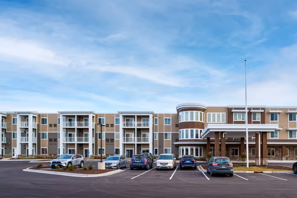 Front exterior of a three-story senior living building with balconies, a rounded main entrance tower and a parking lot under a blue sky.