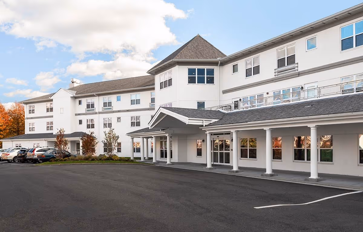 Exterior view of a large, white senior living facility building with multiple windows and a covered entrance supported by white columns. Several cars are parked in the parking lot in front of the building, and trees with autumn foliage are visible in the background under a partly cloudy sky.