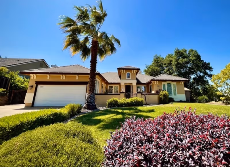 A single-story residential building with a beige exterior, a two-car garage, and a well-maintained front yard featuring green shrubs, a palm tree, and purple-leaved plants under a clear blue sky.