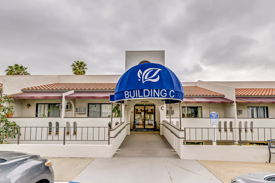 Exterior view of Building C at Rancho Vista Senior Living, featuring a blue awning with white text and logo above the entrance. The building has a beige facade with a tiled roof, windows with air conditioning units, and a ramp leading to the entrance. There are palm trees visible in the background under a cloudy sky.