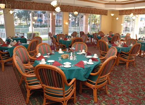 A spacious dining room with multiple round tables covered with green tablecloths and set with white cups, saucers, and folded burgundy napkins. The chairs have wooden frames with striped cushions. Large windows let in natural light and offer a view of the outdoor area with trees and buildings.