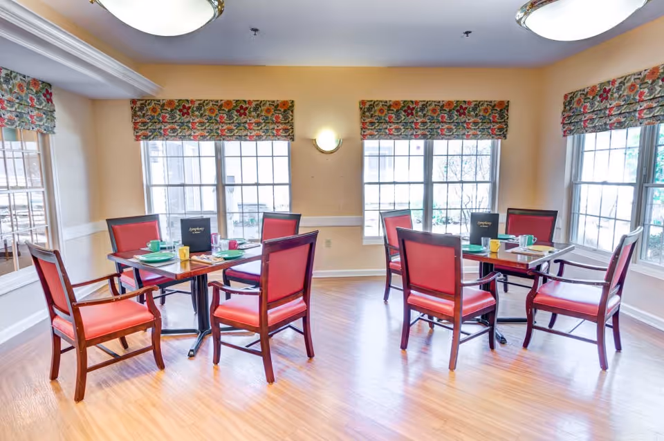 A bright dining room with two square wooden tables, each surrounded by four red cushioned chairs. The tables are set with colorful cups, plates, and menus. Large windows with floral-patterned valances let in natural light, and the room has light-colored walls and wood flooring.