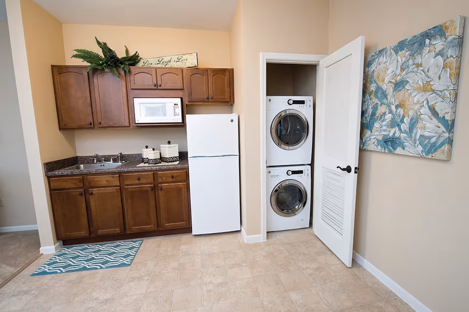 A compact kitchen area with wooden cabinets, a white refrigerator, a microwave, and a sink. Next to the kitchen is a small laundry closet with a stacked washer and dryer behind a partially open white door. The walls are beige, and there is a decorative blue and white floral painting on the right wall. A small green plant and a sign that reads 'Live, Laugh, Love' are placed above the cabinets.