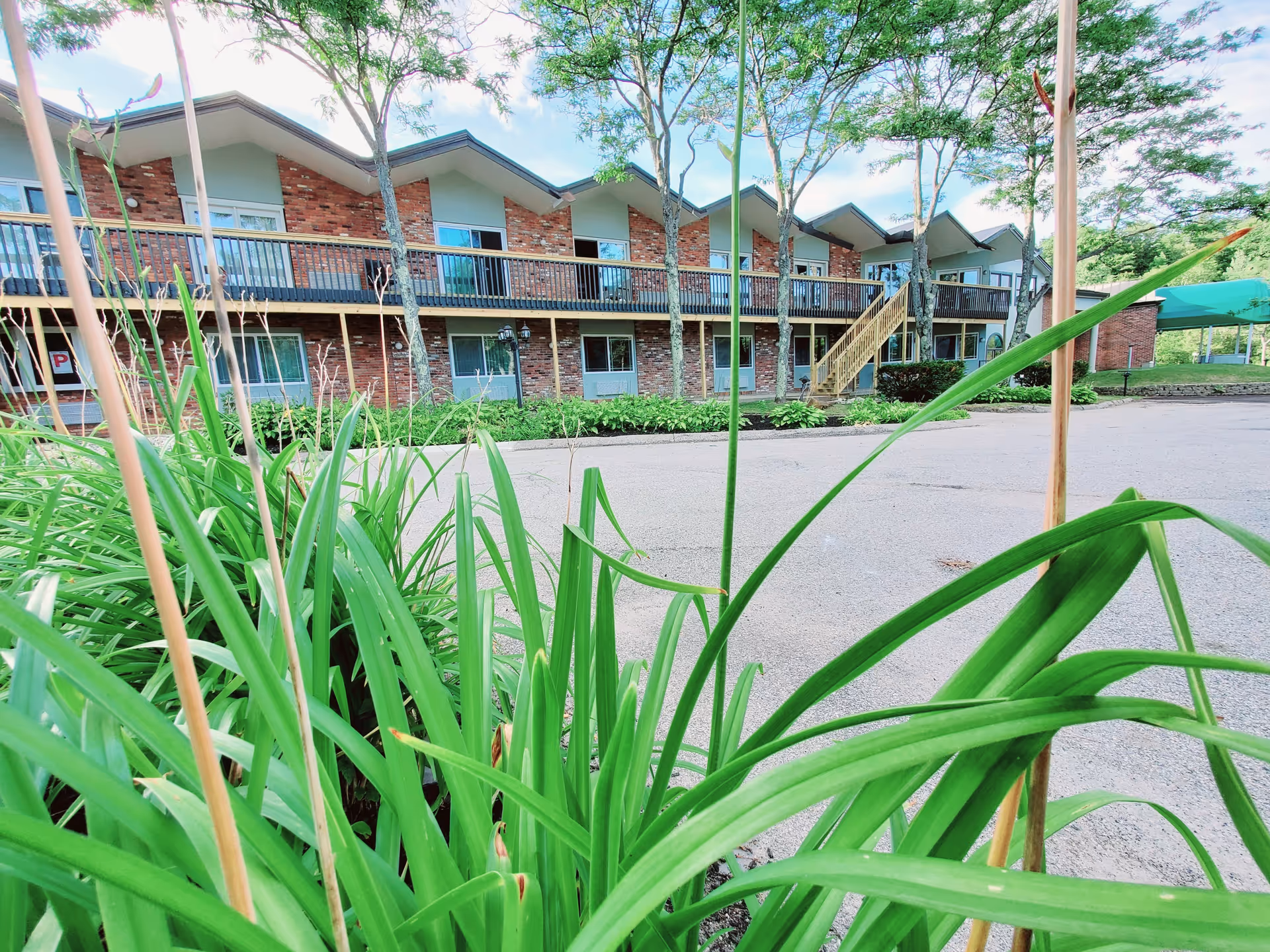 View of a two-story senior living facility building with a brick facade and a balcony running along the upper floor. There are several windows and doors visible, with a staircase leading to the upper level. In the foreground, there are green plants and trees, and a paved driveway or parking area in front of the building under a partly cloudy sky.