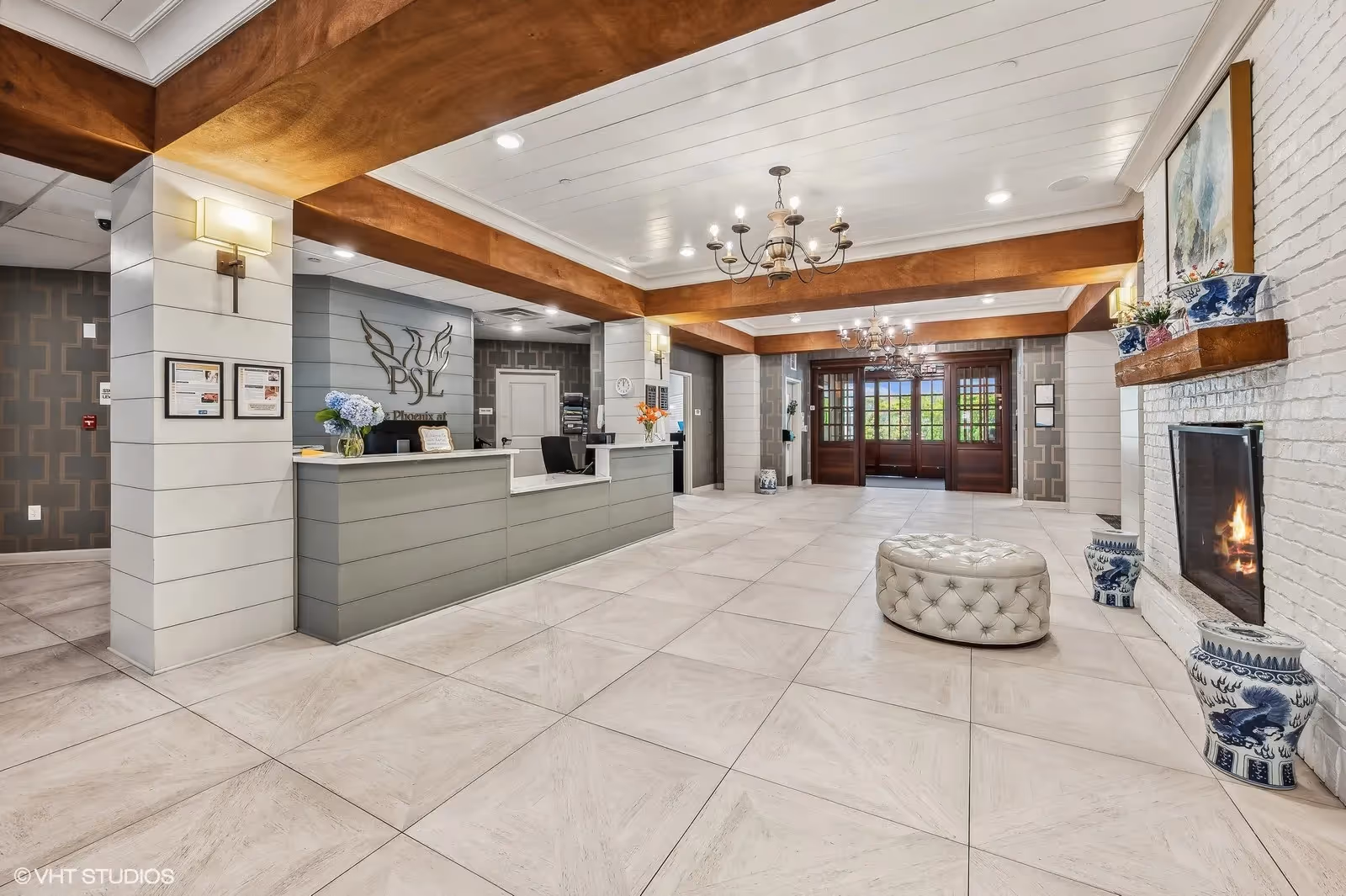 Spacious reception lobby with a front desk, chandeliers, a fireplace, tufted ottoman and decorative vases.
