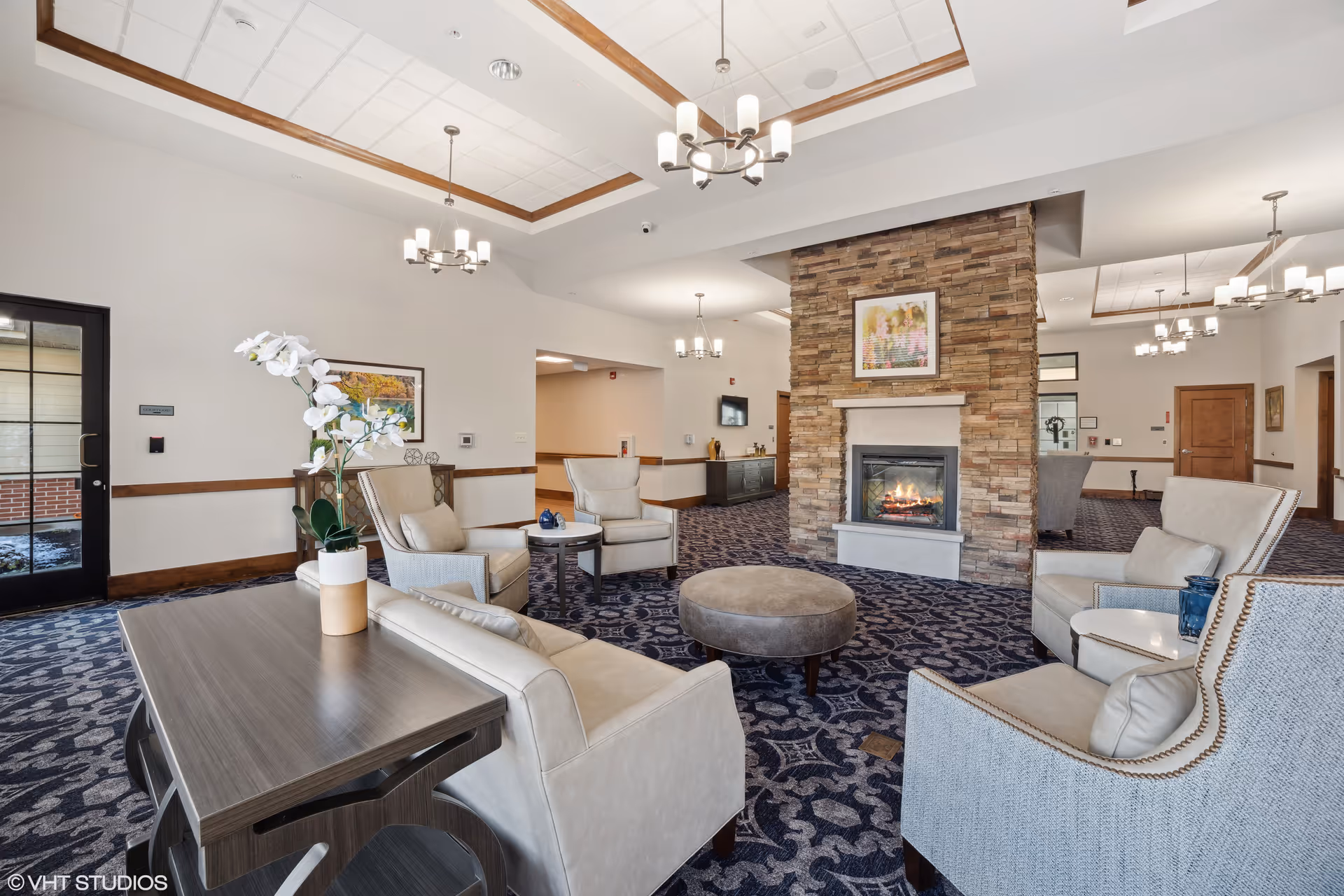 A spacious and well-lit living room area in AVIVA Merrillville featuring comfortable beige armchairs and a sofa arranged around a round ottoman. A stone fireplace with a framed picture above it is centrally located, providing a cozy ambiance. The room has patterned blue carpet, white walls with wooden trim, multiple modern chandeliers, and a table with a white orchid in a pot. Doors and hallways lead to other parts of the facility.